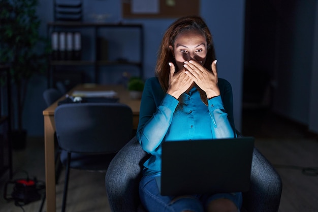 Donkerbruine vrouw die 's nachts op kantoor werkt, bedekt mond met handen voor fout geheim concept