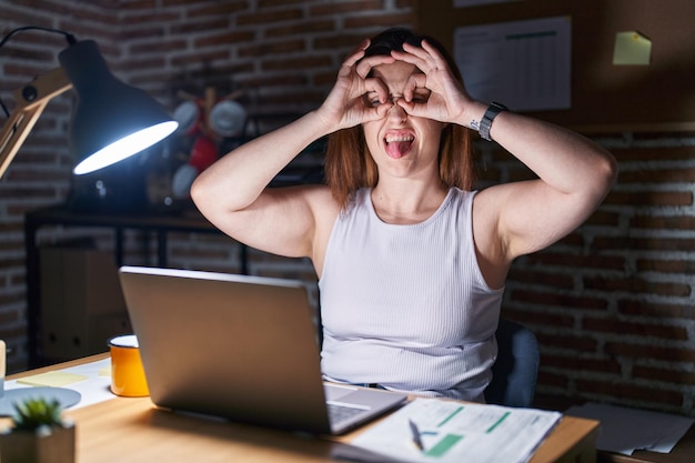 Gratis foto donkerbruine vrouw die 's nachts op kantoor werkt en een goed gebaar doet, zoals een verrekijker die de tong uit de ogen steekt en door de gekke uitdrukking van de vingers kijkt