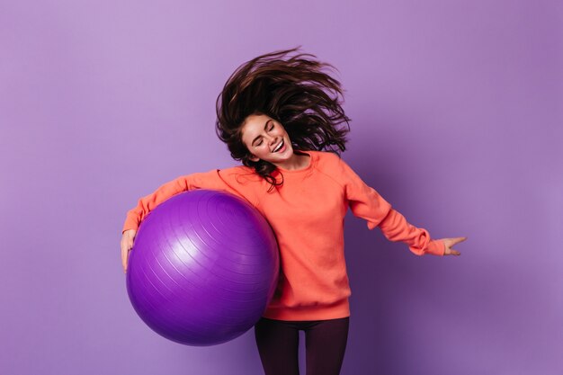 Donkerbruine vrouw in oranje sweatshirt speelt met haar en houdt paarse fitball op geïsoleerde muur