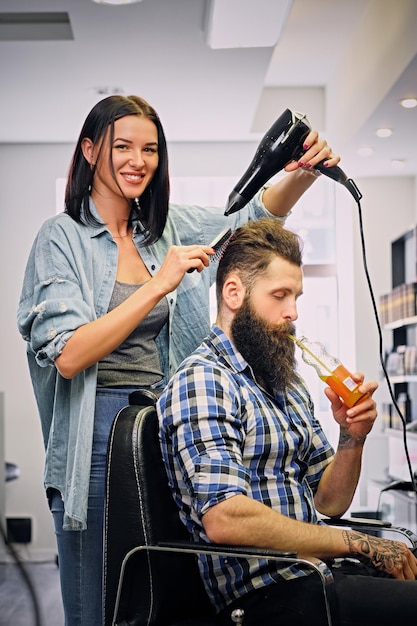 Donkerbruine vrouwelijke kapper die een kapsel met föhn van de bebaarde mannencliënt in een salon doet.