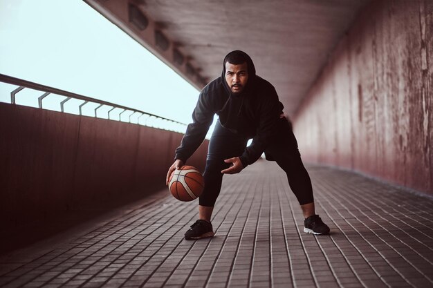 Donkere man gekleed in een zwarte hoodie en sportshort die basketbal speelt op een voetpad onder een brug, kijkend naar een camera.