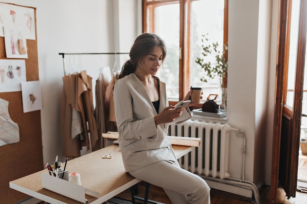 Gratis foto donkerharige dame in wit pak met koffie en telefoon charmante vrouw in linnen broek en beige blouse chatten in mobiele telefoon en thee drinken