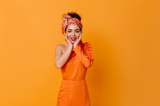 Donkerharige dame met rode lippenstift gekleed in oranje jurk en Aheadband met glimlach camera kijken op geïsoleerde ruimte.