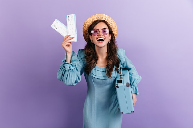 Donkerharige meisje met bril en strooien hoed houdt kaartjes en blauwe koffer. Portret van reiziger in schattige retro jurk op lila muur.
