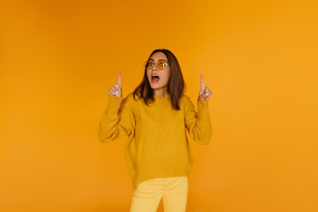 Donkerharige mooi meisje in glazen poseren op gele muur. Trendy Europese vrouw die verbazing uitdrukt.