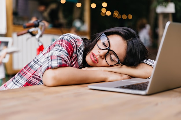 Donkerharige mooie student slapen in de buurt van computer. Outdoor Portret van charmante vrouwelijke freelancer in geruit overhemd restng na het werk.