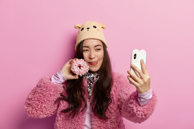 Donkerharige tiener likt lippen, poseert met heerlijke zoete geglazuurde donut, keert terug uit bakkerijwinkel, neemt selfie op mobiele telefoon, heeft plezier