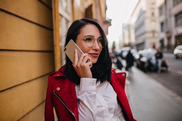 Donkerharige vrouw in wit overhemd en rode jas praten over de telefoon op stadsmuur