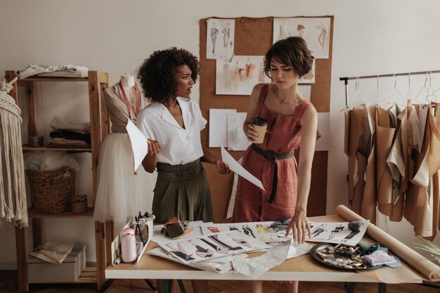 Donkerhuidige jonge krullende vrouw in kaki korte broek en witte blouse houdt vellen papier vast en praat met kortharig meisje in linnen jurk Vrouwen werken als modeontwerpers en zien er ontevreden uit