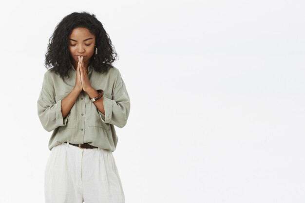 Donkerhuidige vrouw die haar hoofd naar beneden buigt, ogen sluit die vredig en ontspannen staan met de handen in bidden