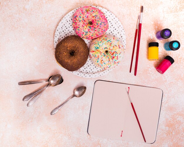 donuts; penseel; lepels en verf fles op lege notebook over de gestructureerde achtergrond