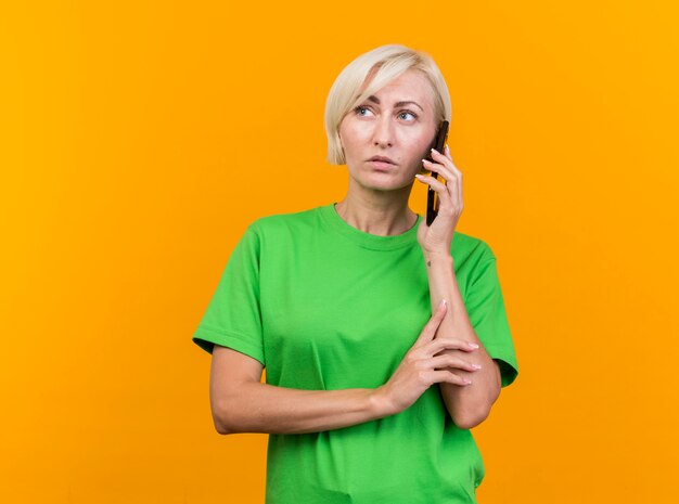 Doordachte blonde Slavische vrouw van middelbare leeftijd praten over de telefoon aanraken van arm kijken kant geïsoleerd op gele achtergrond met kopie ruimte