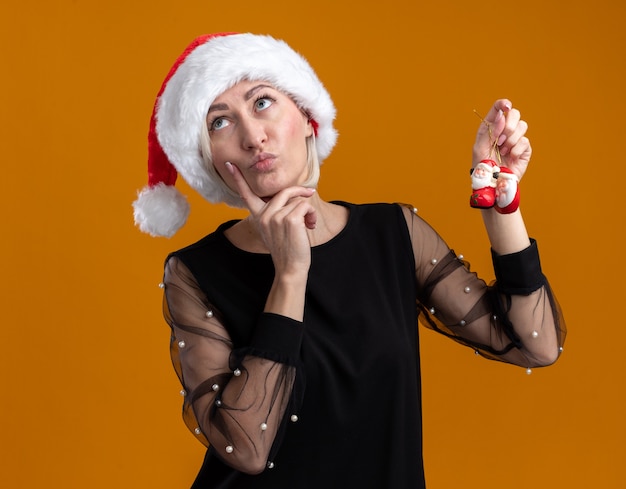 Doordachte blonde vrouw van middelbare leeftijd met kerstmuts opzoeken met kerstman kerst ornamenten aanraken kin geïsoleerd op een oranje achtergrond