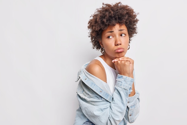 Doordachte ellendige neerslachtige krullende Afro-Amerikaanse vrouw houdt handen onder kin portemonnees lippen kijkt droevig weg draagt stijlvolle outfit poses tegen witte muur