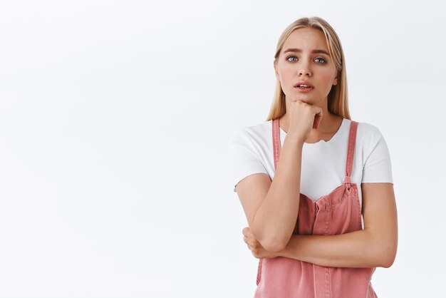 Doordachte en verontruste jonge bezorgde blonde vrouw die aan iets denkt over hand onder de kin vasthoudend fronsend staren bezorgd als nadenkend over belangrijke beslissing staan witte achtergrond