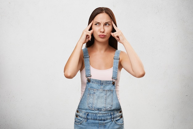 Doordachte geconcentreerde vrouw in denim overall, vingers op tempels houden, proberen idee te krijgen, peinzend opzij kijken, geïsoleerd over witte blinde muur. Jonge vrouw probeert terug te roepen juiste antwoord