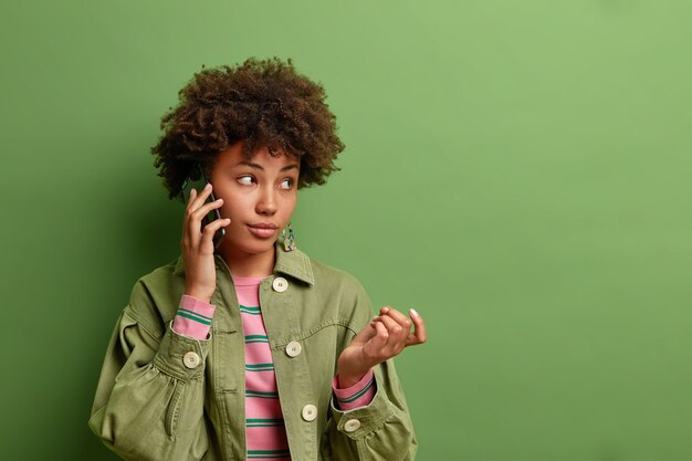 Doordachte jonge Afro-Amerikaanse vrouw steekt hand op heeft telefoongesprek overweegt informatie gehoord van gesprekspartner draagt stijlvolle kleding geïsoleerd over levendige groene muur