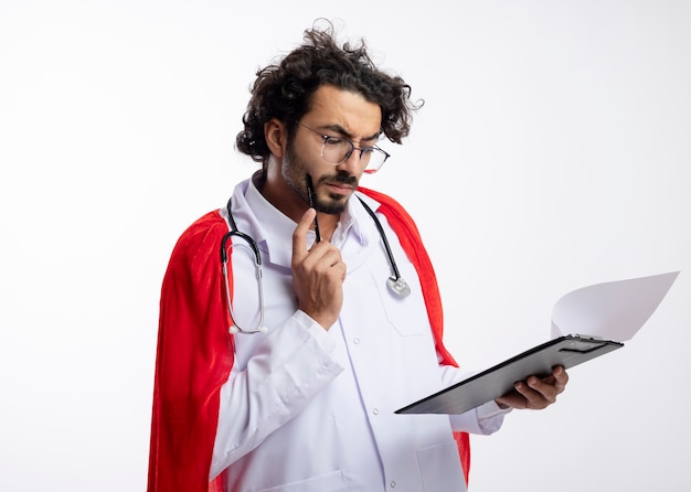 Doordachte jonge blanke man in optische bril dragen arts uniform met rode mantel en met een stethoscoop om de nek zet potlood op gezicht en kijkt naar Klembord met kopie ruimte