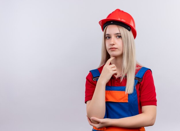 Doordachte jonge blonde ingenieur bouwer meisje in uniform vinger op kin zetten geïsoleerde witte muur met kopie ruimte