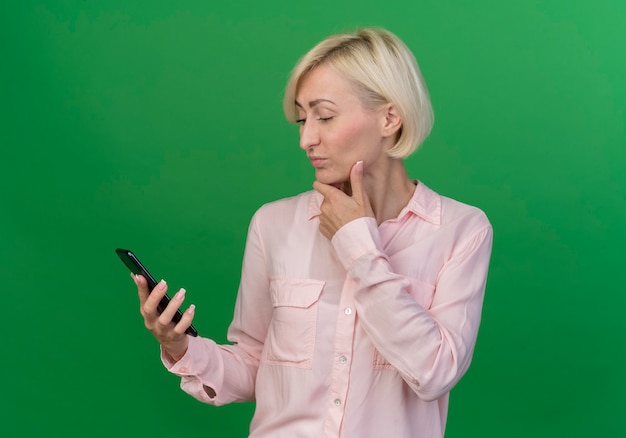 Gratis foto doordachte jonge blonde slavische vrouw houden en kijken naar mobiele telefoon en kin geïsoleerd op groene achtergrond aan te raken