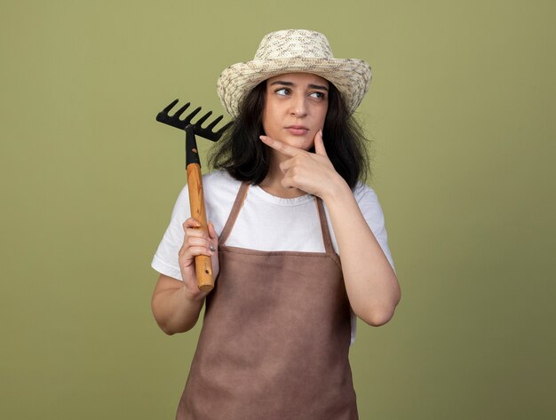 Doordachte jonge brunette vrouwelijke tuinman in uniform dragen tuinieren hoed legt hand op kin en houdt hark kijken kant geïsoleerd op olijfgroene muur