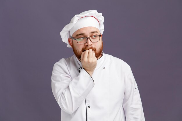 Doordachte jonge chef-kok met een uniforme bril en pet kijkend naar bijtende vingers geïsoleerd op paarse achtergrond