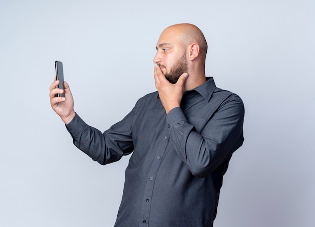 Doordachte jonge kale call center man houden en kijken naar mobiele telefoon met hand op mond geïsoleerd op een witte muur