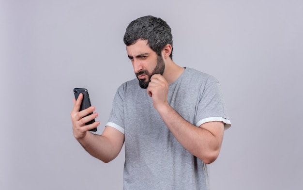 Doordachte jonge knappe man houden en kijken naar mobiele telefoon en zijn kin aan te raken geïsoleerd op een witte muur