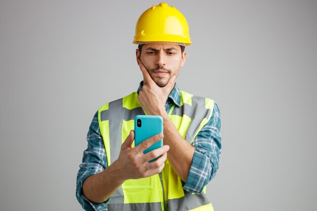 Doordachte jonge mannelijke ingenieur die een veiligheidshelm en uniform draagt en naar mobiele telefoon kijkt terwijl hij de hand op de kin houdt, geïsoleerd op een witte achtergrond