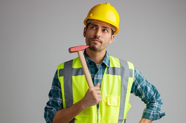 Doordachte jonge mannelijke ingenieur met veiligheidshelm en uniform aanraken kin met hamer opzoeken met hand op taille geïsoleerd op witte achtergrond