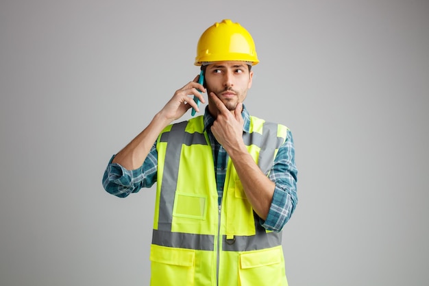 Doordachte jonge mannelijke ingenieur met veiligheidshelm en uniform kijkend naar de zijkant die de hand op de kin houdt terwijl hij aan de telefoon praat geïsoleerd op een witte achtergrond