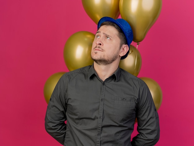 doordachte jonge partij kerel met blauwe hoed achter ballonnen geïsoleerd op roze