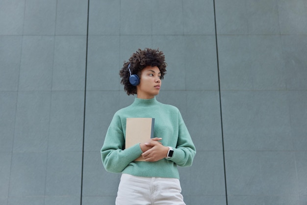 Gratis foto doordachte jonge vrouw met krullend haar houdt notitieblok luistert audiotrack via koptelefoon draagt casual trui en witte spijkerbroek gefocust weg poses tegen grijze muur lege ruimte voor uw promotie