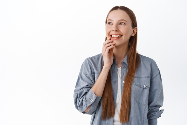 Doordachte jonge vrouw met lang haar die denkt, tevreden glimlacht en opzij kijkt naar de promotietekst van de kopieerruimte, iets in beeld brengt, staande tegen een witte muur