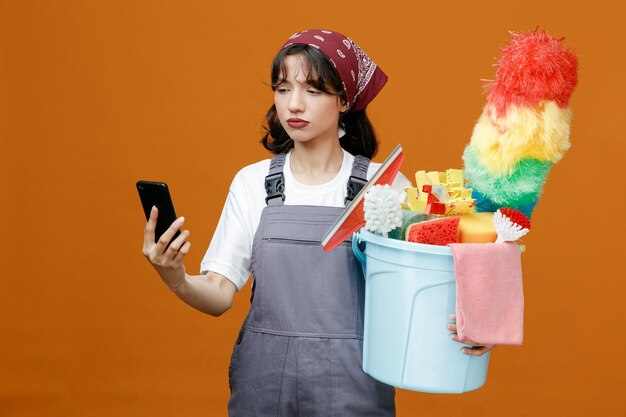 Doordachte jonge vrouwelijke schoonmaker met uniform en bandana met mobiele telefoon en emmer met schoonmaakhulpmiddelen kijkend naar mobiele telefoon geïsoleerd op oranje achtergrond
