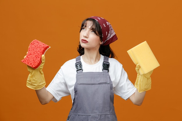 Doordachte jonge vrouwelijke schoonmaker met uniforme rubberen handschoenen en bandana kijkend naar camera met sponzen geïsoleerd op oranje achtergrond