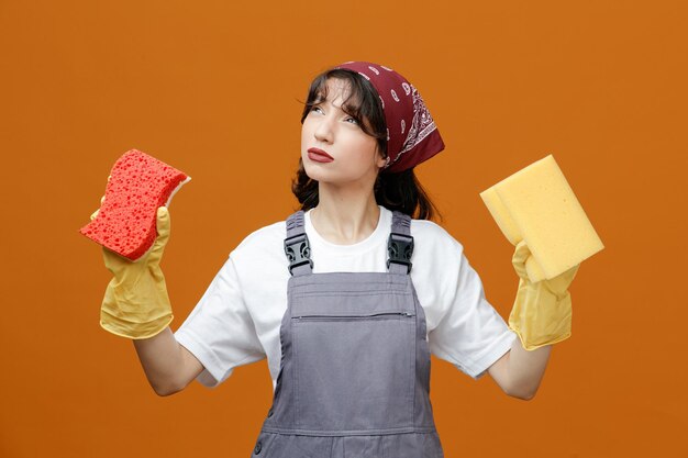 Doordachte jonge vrouwelijke schoonmaker met uniforme rubberen handschoenen en bandana kijkend naar camera met sponzen geïsoleerd op oranje achtergrond