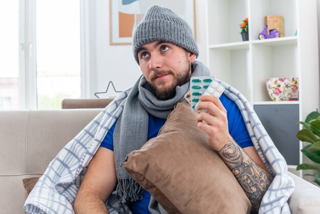 Doordachte jonge zieke man met sjaal en wintermuts gewikkeld in deken zittend op de bank in de woonkamer met kussen en pakjes pillen omhoog kijkend