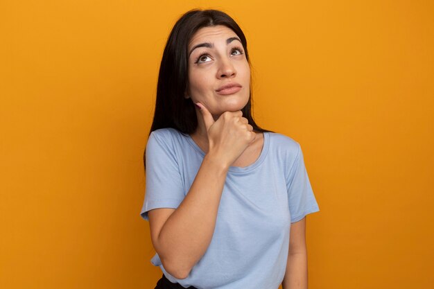 Doordachte mooie brunette vrouw legt hand op kin en kijkt omhoog geïsoleerd op oranje muur