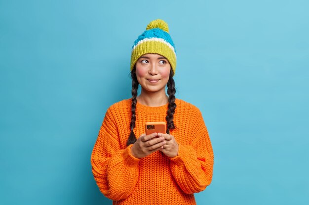 Doordachte mooie tienermeisje houdt mobiele telefoon vast en stuurt sms-berichten denkt welk antwoord te geven leest opmerkingen onder haar bericht in sociale netwerken gekleed in winterkleren geïsoleerd op blauwe muur