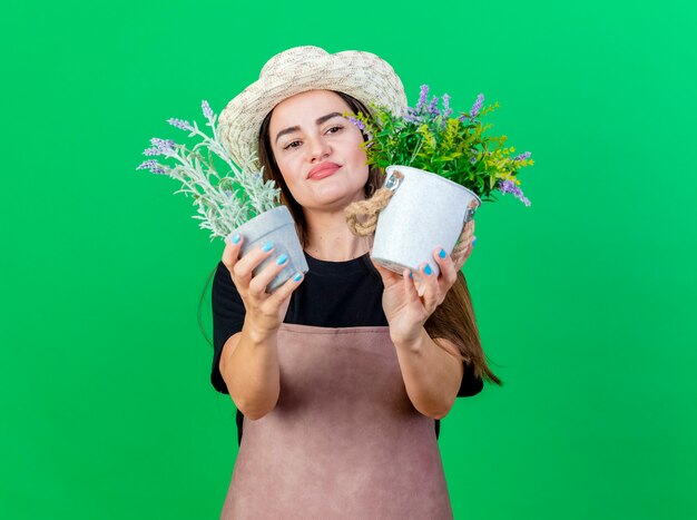 doordachte mooie tuinman meisje in uniform dragen tuinieren hoed bedrijf en kijken naar bloem in bloempot geïsoleerd op groen