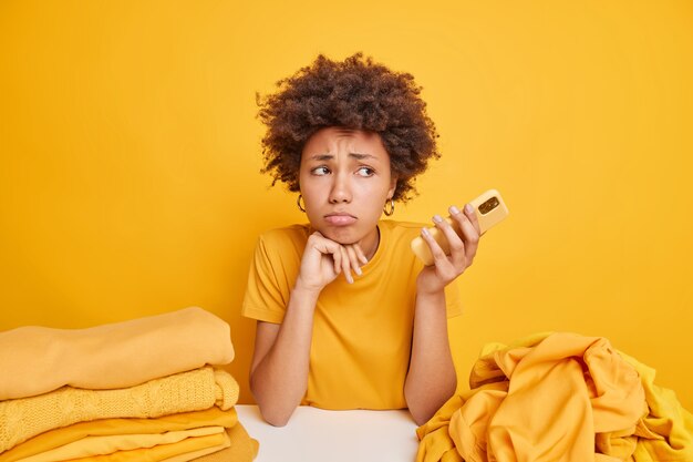 Doordachte ontevreden Afro-Amerikaanse vrouw houdt smartphone vast en wacht op telefoontje voelt zich moe na het doen van huishoudelijke klusjes vouwt de was op na het wassen poses aan tafel met stapels gewassen kleren in de buurt