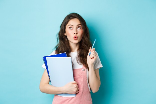 Doordachte slimme vrouwelijke student die een pen vasthoudt en een idee heeft, peinzend opkijkt, notitieboekjes draagt, staande tegen een blauwe achtergrond.
