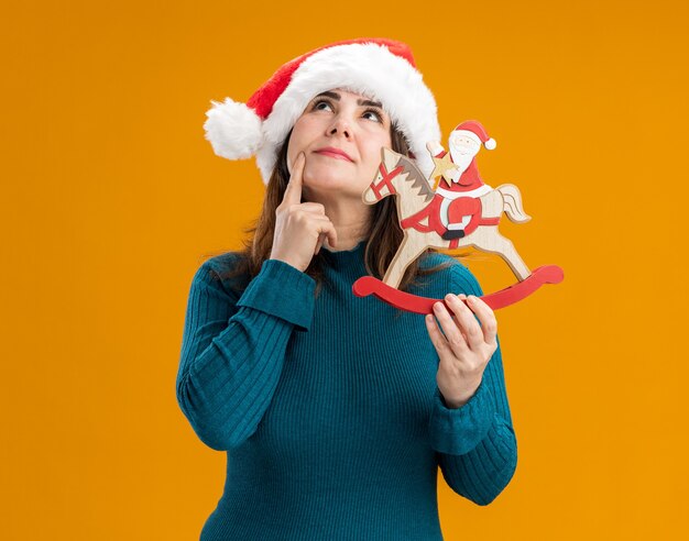 Doordachte volwassen blanke vrouw met kerstmuts legt vinger op kin en houdt santa op schommelpaard decoratie opzoeken geïsoleerd op een oranje achtergrond met kopie ruimte