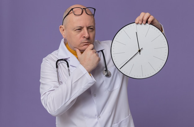 Doordachte volwassen Slavische man met bril in doktersuniform met stethoscoop die klok vasthoudt