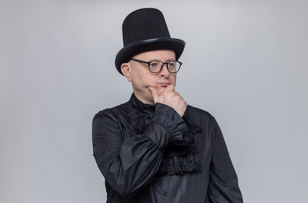 Doordachte volwassen Slavische man met hoge hoed en optische bril in zwart gotisch shirt die hand op zijn kin legt en naar de zijkant kijkt