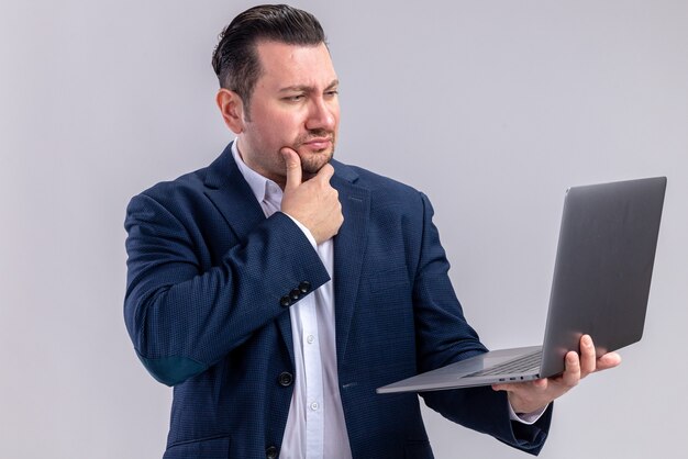 Doordachte volwassen Slavische zakenman die laptop vasthoudt en bekijkt die op een witte muur met kopieerruimte wordt geïsoleerd