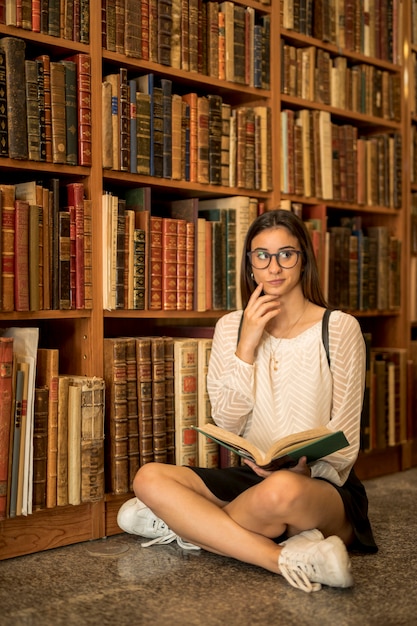 Doordachte vrouwelijke studentenzitting in kleermakerszit met boek in bibliotheek