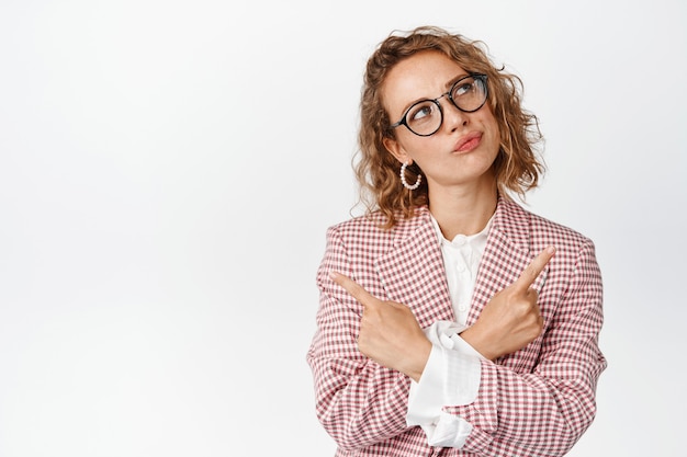 Doordachte zakenvrouw met een bril en een stijlvol pak, die er geïntrigeerd uitziet en zijwaarts wijst, beslissingen neemt, denkt, kiest uit varianten op wit.