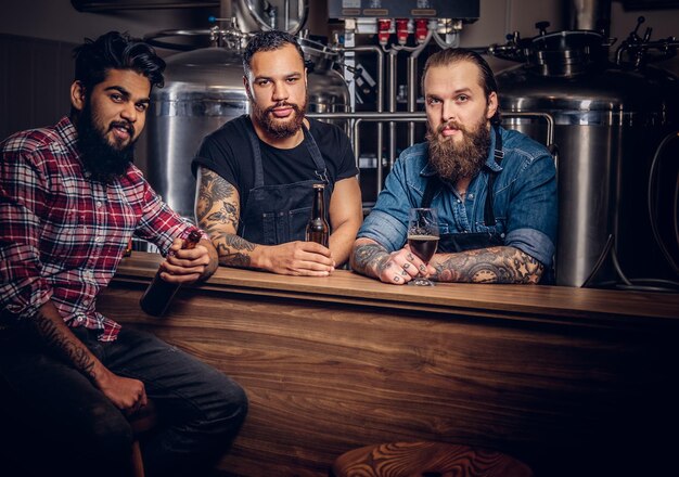 Drie bebaarde interraciale vrienden drinken ambachtelijk bier in een brouwerij. Twee hipsterarbeiders in schorten drinken bier met hun vriend in een brouwerijfabriek.